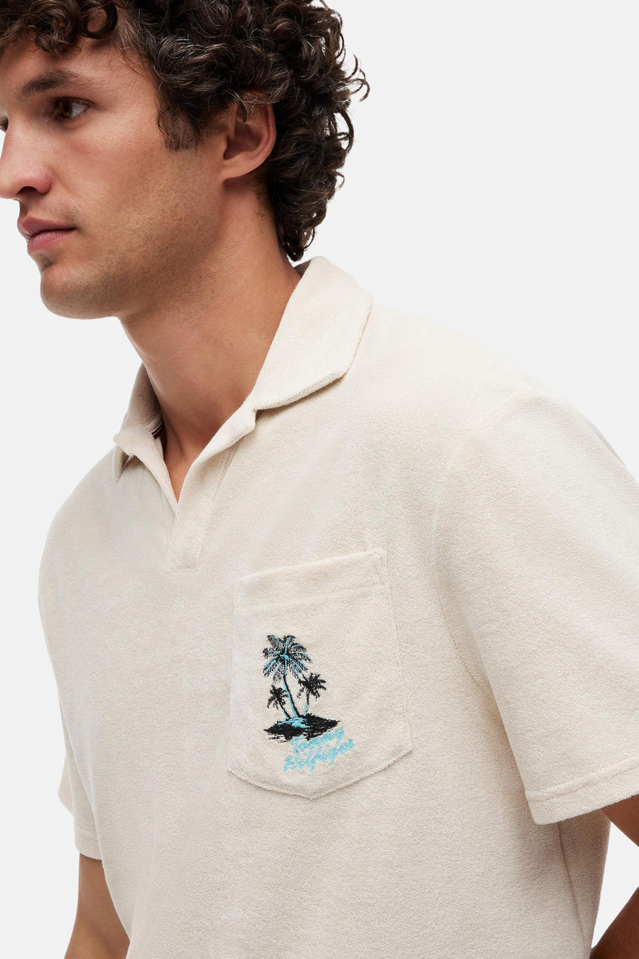 Polo écru en tissu bouclette de TOMMY HILFIGER avec motif palmiers et script sur la poche poitrine, porté par un homme.