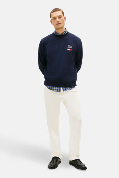 Sweat bleu de TOMMY HILFIGER, porté sur une chemise à carreaux et un jean blanc avec des chaussures noires.