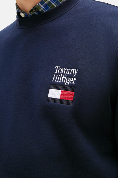 Sweat bleu de TOMMY HILFIGER avec logo brodé, porté sur une chemise à carreaux.
