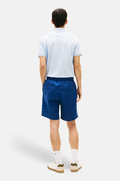 Bermuda bleu de TOMMY HILFIGER avec poches boutonnées à l'arrière, porté avec un polo bleu clair et des baskets blanches.
