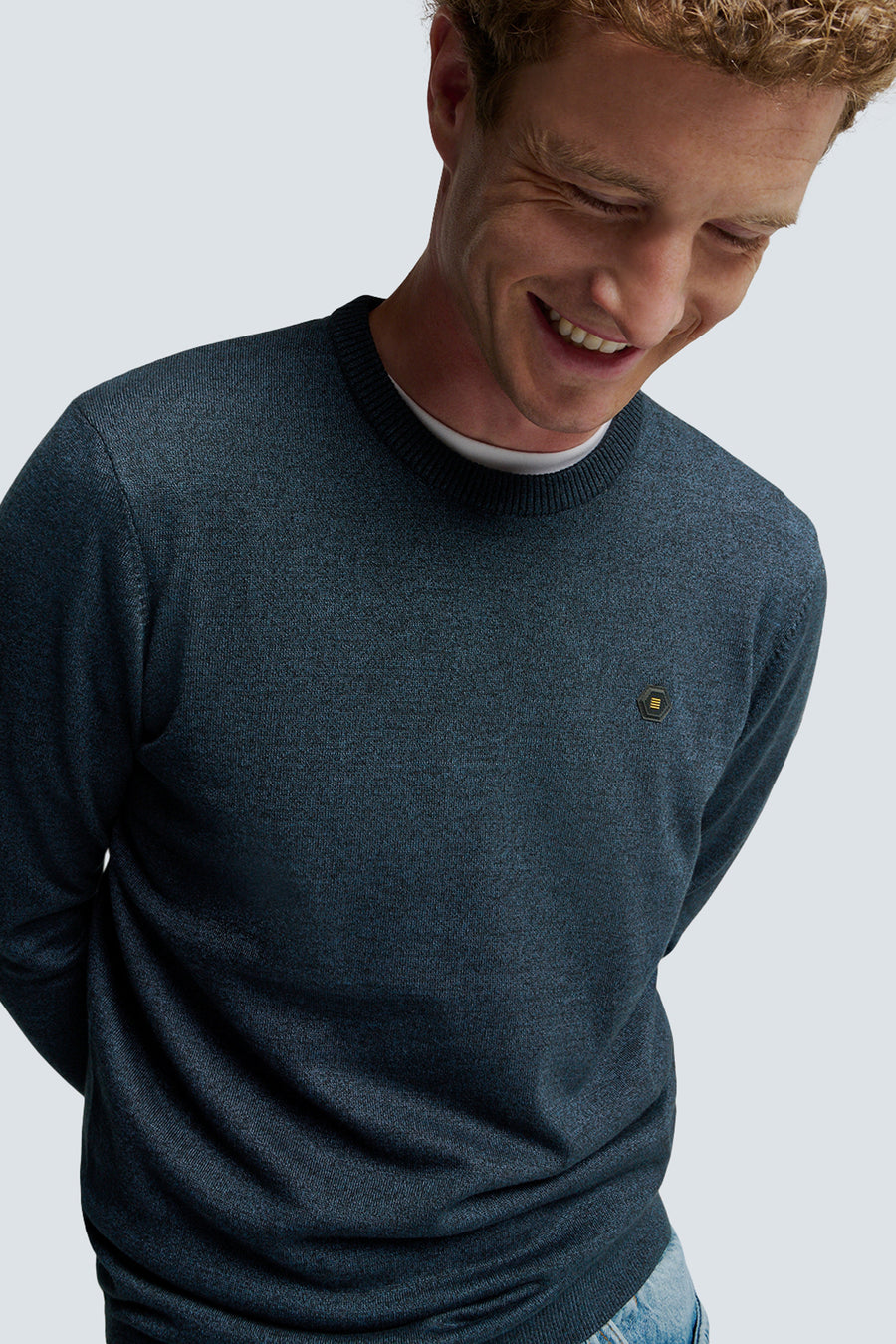 Pull à col rond bleu chiné de NO EXCESS, avec écusson à motifs géométriques, porté avec un t-shirt blanc.
