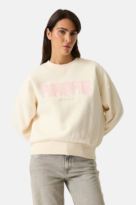 Sweat-shirt écru de GARCIA, avec le mot « amore » rose en relief sur le devant, porté avec un jean.

