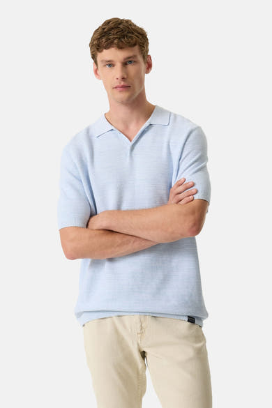 Polo en tricot bleu clair de GARCIA, porté avec un pantalon beige, bras croisés.
