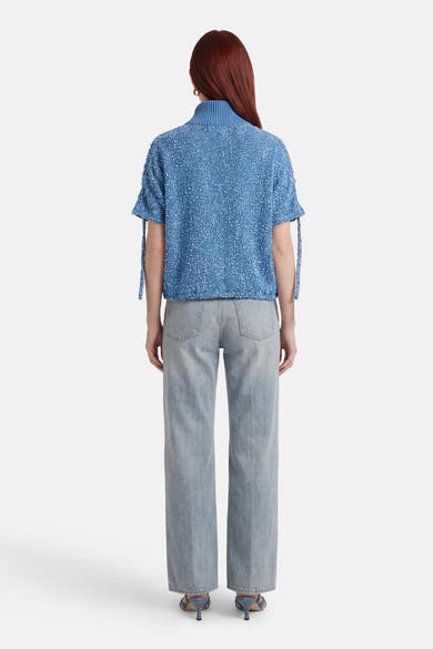 Veste courte bleue texturée de ADORE, avec des liens décoratifs aux manches, portée avec un jean et des chaussures assorties.