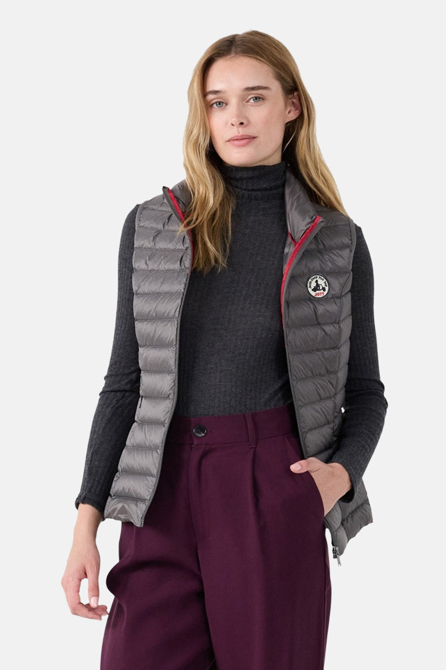 Veste sans manches grise de JOTT, avec doublure rouge, portée sur un pull gris foncé et un pantalon bordeaux.