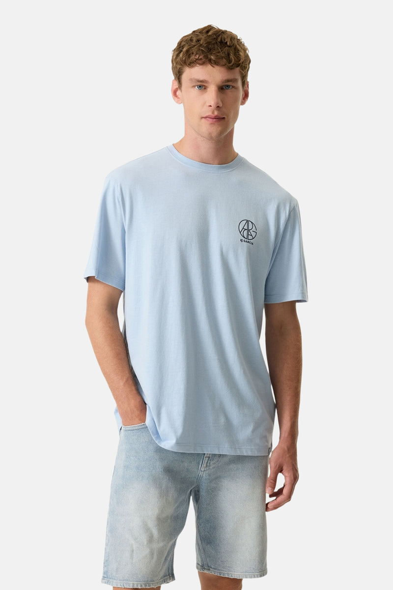 Lichtblauwe T-shirt van GARCIA met een zwart cirkelvormig "AB GARCIA" logo op de borst, gedragen door een man in een lichtblauwe spijkerbroek.