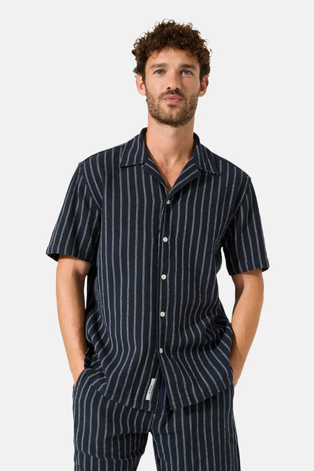 Chemise bleu marine à rayures de GARCIA, à manches courtes et col cubain, portée avec un pantalon assorti.