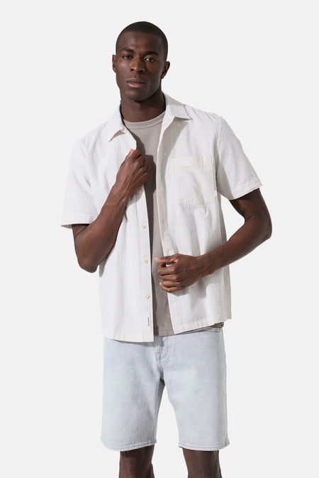 Chemise à manches courtes beige de GARCIA, texturée à fines rayures verticales, portée ouverte sur un t-shirt gris et un short en jean clair.