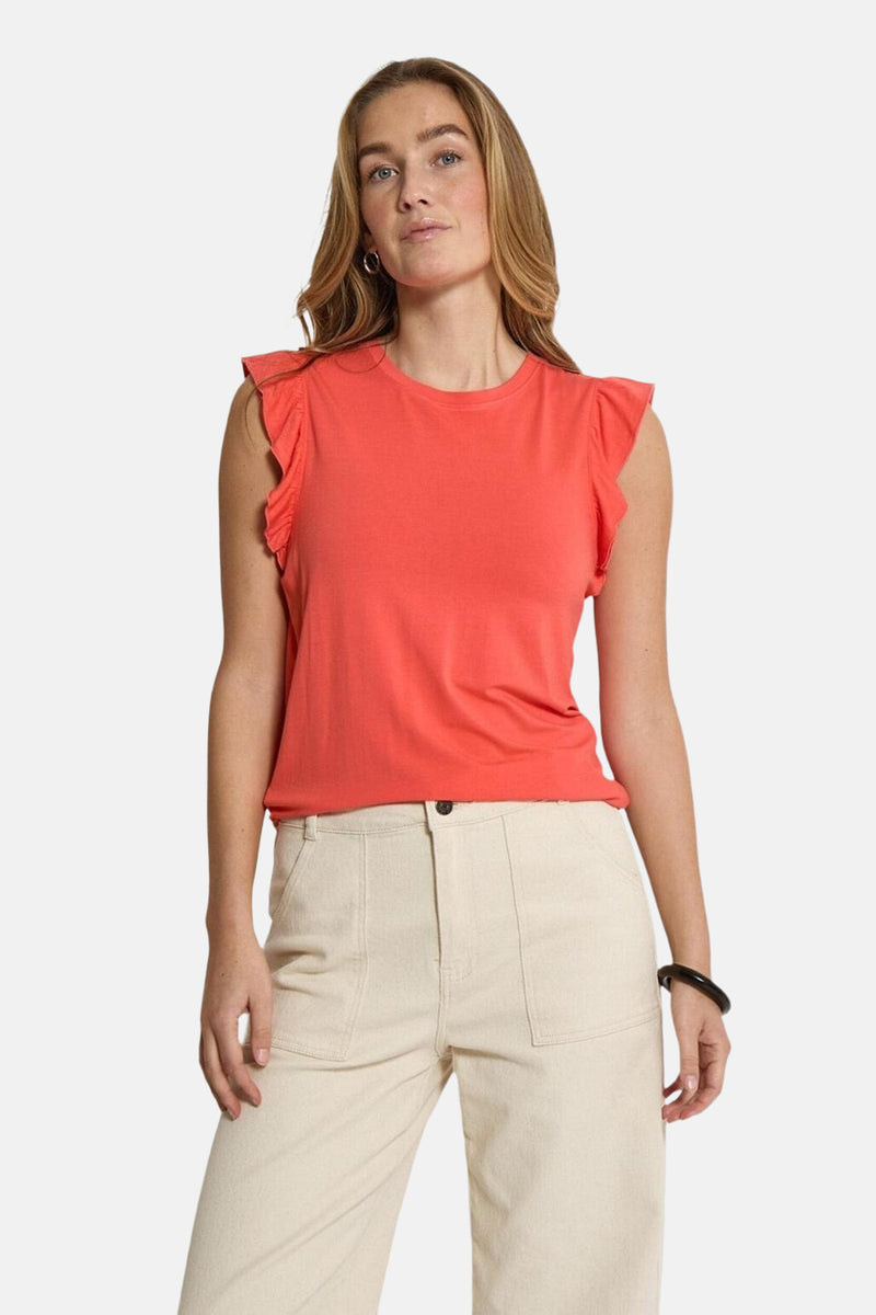 Top sans manches corail de PEPPERCORN, avec des volants aux épaules et porté avec un pantalon beige.
