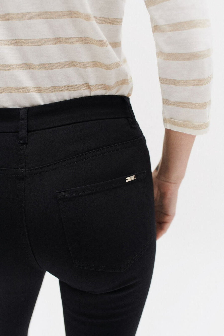 Zwarte broek van CAROLL, nauwsluitende pasvorm, achterzakdetail met metalen logo, gedragen met een gestreept topje.