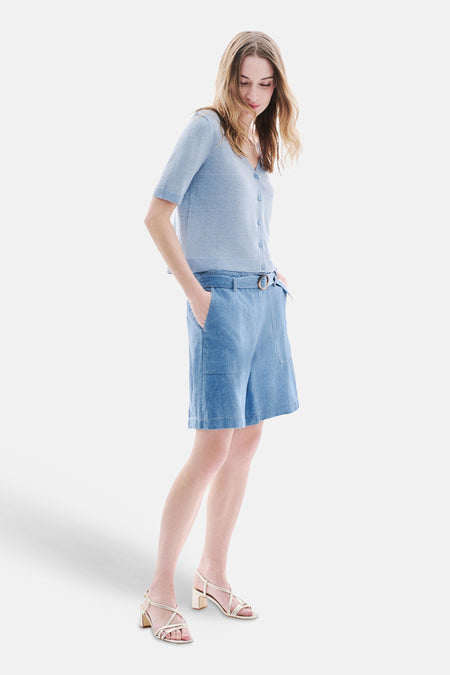Short en jean bleu clair de CAROLL avec ceinture à boucle ronde et poches, porté avec un cardigan bleu et des sandales dorées.