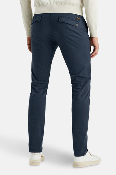 Blauwe chino van PME Legend, met knoopbare achterzakken, gedragen met een lichtgekleurde trui en witte schoenen.