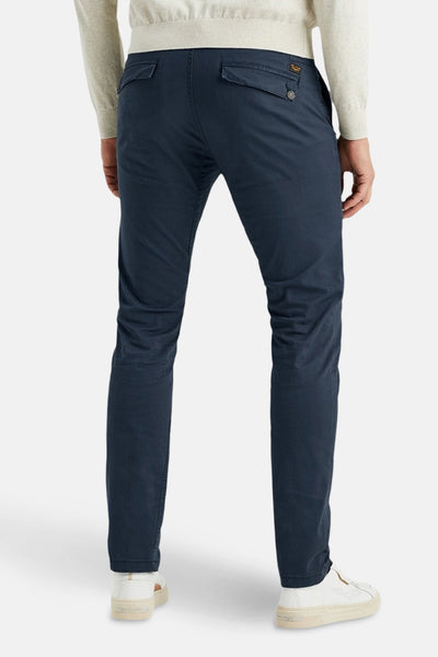 Blauwe chino van PME Legend, met knoopbare achterzakken, gedragen met een lichtgekleurde trui en witte schoenen.