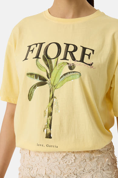 T-shirt jaune de GARCIA avec imprimé palmier banane, texte "FIORE" et "love, Garcia", porté avec une jupe en dentelle.