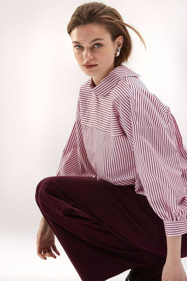 Pink striped long-sleeve blouse with relaxed fit and contrast panel, worn by model in side view with burgundy pants.