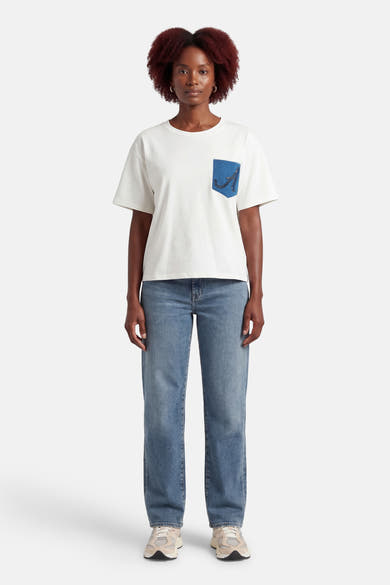 T-shirt blanc à manches courtes de ADORE, avec une poche bleue décorée, porté avec un jean et des baskets.
