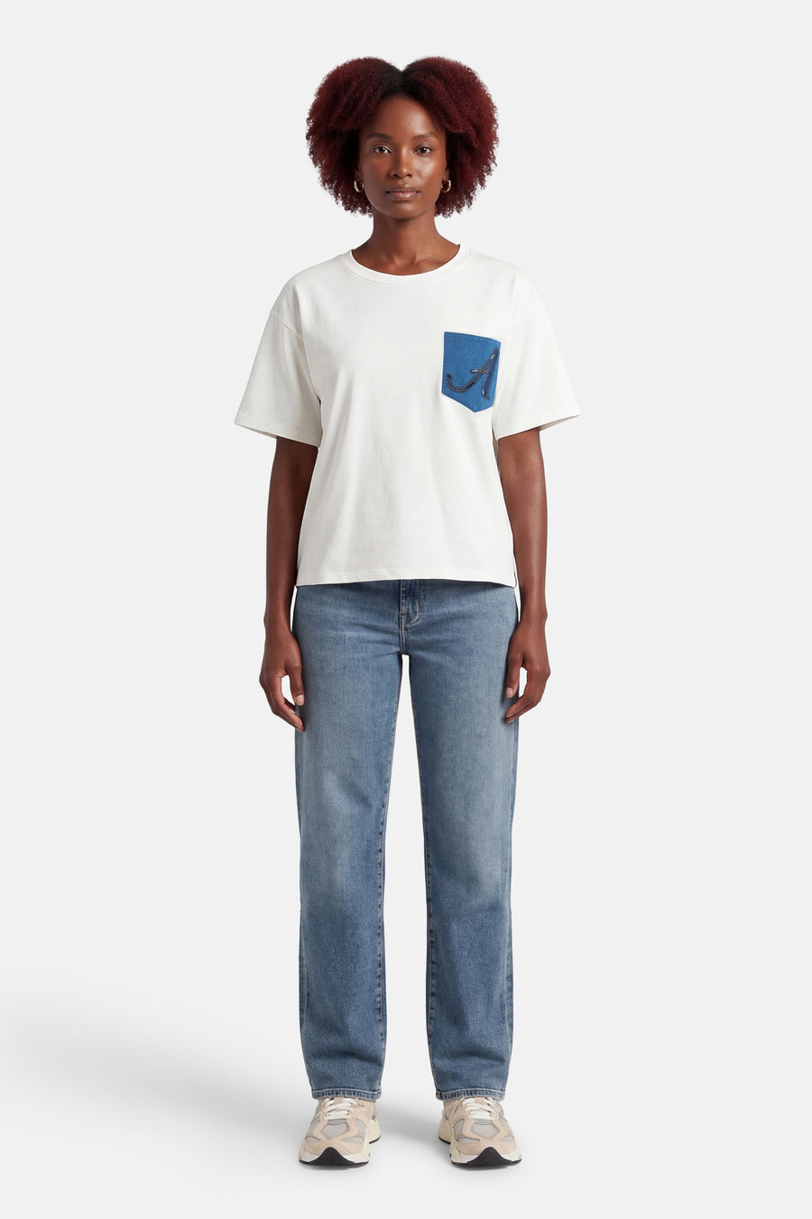 T-shirt blanc à manches courtes de ADORE, avec une poche bleue décorée, porté avec un jean et des baskets.
