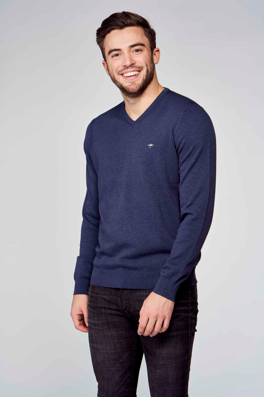 Pull col V bleu de Fynch-Hatton, porté avec un pantalon noir texturé, homme souriant sur fond uni.
