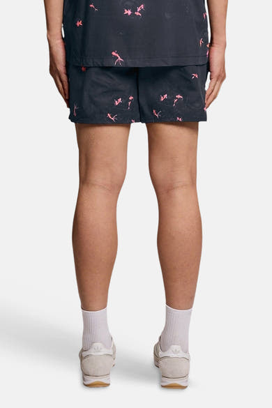 Short de bain bleu de LYLE & SCOTT, à motif de poissons roses, porté de dos avec un haut assorti, chaussettes et baskets claires.