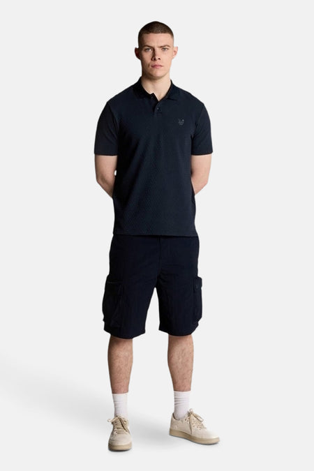 Blauw poloshirt met korte mouwen van LYLE & SCOTT, gedragen met een marineblauwe cargoshort, witte sokken en crèmekleurige sneakers.