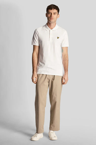 White short-sleeve polo shirt with small yellow bird emblem on chest, worn by model in beige pants and white sneakers, front view.