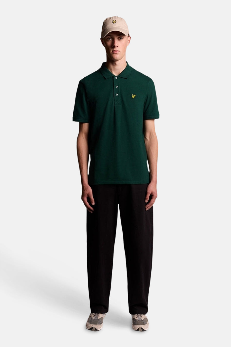 Groen poloshirt van LYLE & SCOTT met klassieke kraag, witte knopen en geel adelaarslogo, gedragen door een man met een beige pet.