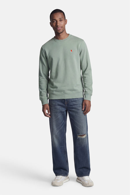 Groene sweater met ronde hals van STROM met rood logo, gedragen door een man in spijkerbroek en sneakers.