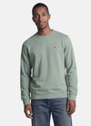 Groene rondehals sweatshirt van STROM met een klein rood-wit geborduurd logo op de borst, gedragen door een man in een spijkerbroek.