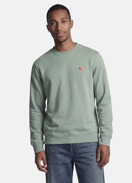 Groene rondehals sweatshirt van STROM met een klein rood-wit geborduurd logo op de borst, gedragen door een man in een spijkerbroek.