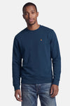 Rondehals sweatshirt STROM blauw met klein oranje en roze geborduurd logo op de borst, gedragen door een man.