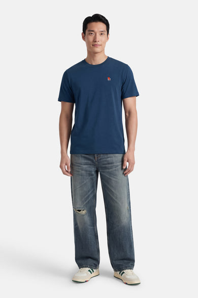 Blauwe T-shirt van STROM met een klein rood logo op de borst, gedragen door een man met een gescheurde spijkerbroek en witgroene sneakers.