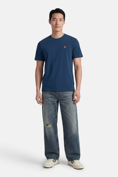 Blauwe T-shirt van STROM met een klein rood logo op de borst, gedragen door een man met een gescheurde spijkerbroek en witgroene sneakers.