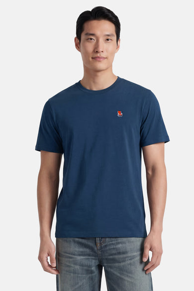 Blauw T-shirt met korte mouwen van STROM, met een klein rood-wit stoellogo geborduurd op de borst, gedragen met een spijkerbroek.