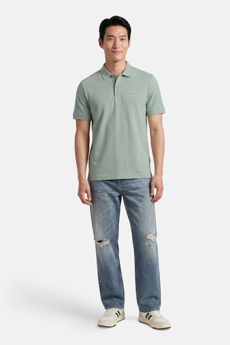 Groen STROM poloshirt met oranje driehoekig logo, gedragen door een man in gescheurde jeans en sneakers.