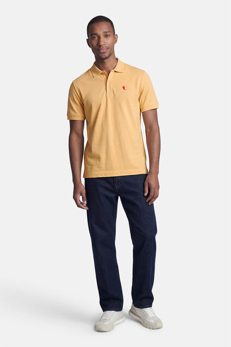 Oranje STROM polo met rood fauteuil logo, gedragen door een man in een blauwe spijkerbroek en lichte sneakers.