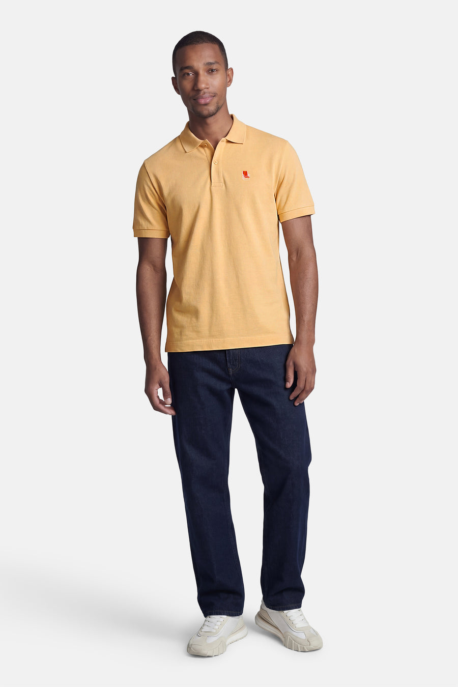 Oranje STROM polo met rood fauteuil logo, gedragen door een man in een blauwe spijkerbroek en lichte sneakers.
