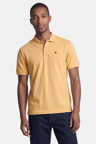 Oranje polo van STROM met een strandstoelborduurwerk op de linkerborst, gedragen door een man in een spijkerbroek.