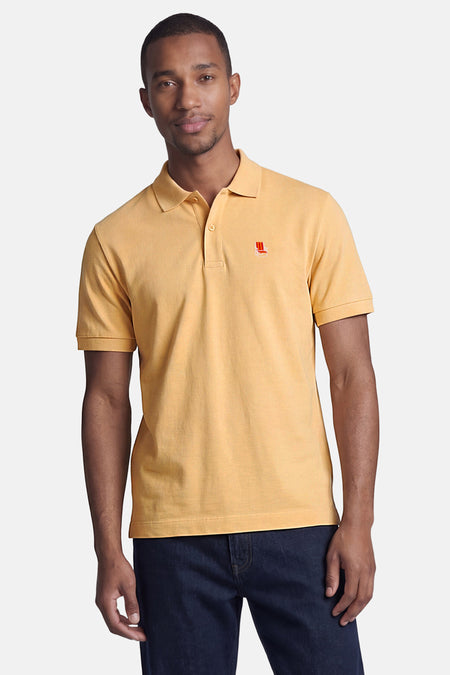 Oranje polo van STROM met een strandstoelborduurwerk op de linkerborst, gedragen door een man in een spijkerbroek.