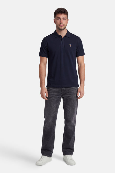 Marineblauwe polo van STROM met een cocktailmotief, gedragen door een man met een grijze jeans en witte sneakers.