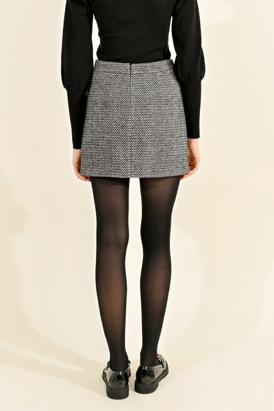 Jupe courte noire Molly Bracken à motif à chevrons, portée avec un pull noir et des collants opaques.