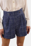 Korte blauwe geruite short van Molly Bracken, van achteren gezien, gedragen met een blouse en rode lakleren enkellaarsjes.