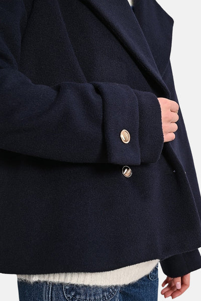 Veste courte bleue par Molly Bracken avec boutons dorés, portée sur un pull blanc et un jean.
