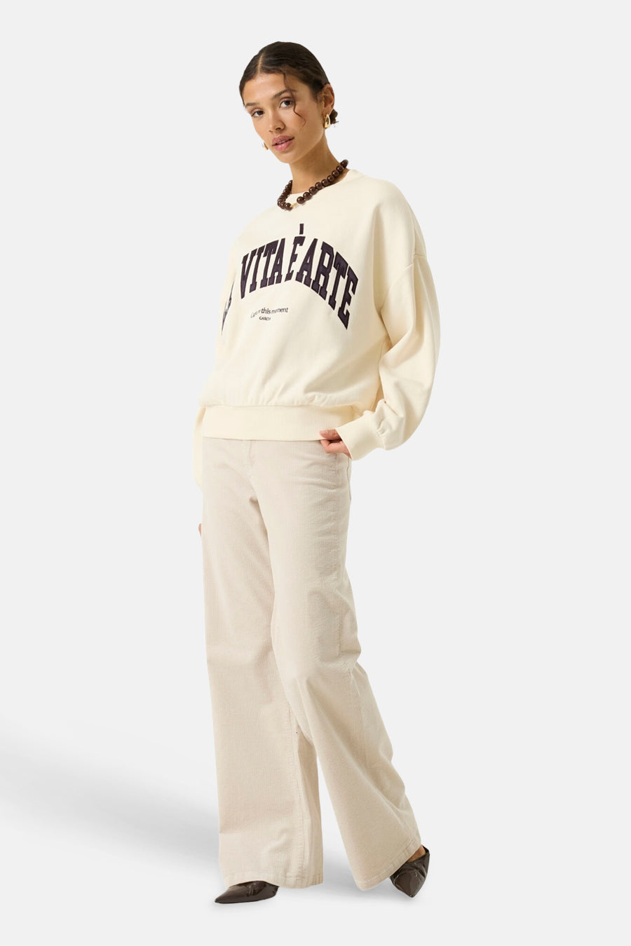 Ecru crewneck sweatshirt with bold black arched text and small tag detail, worn with matching wide-leg corduroy pants and dark brown loafers, side view.