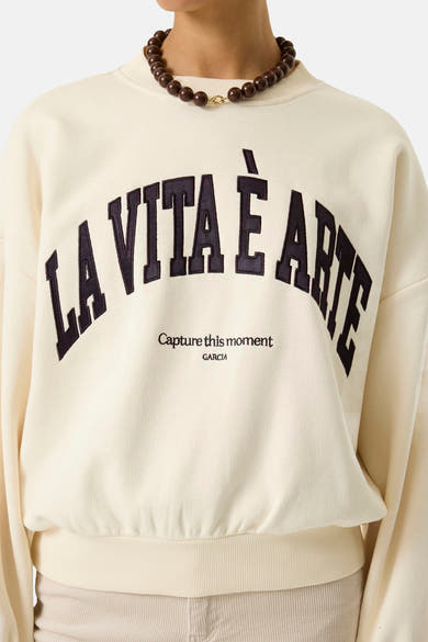 Ecru crewneck sweatshirt with bold black "LA VITA È ARTE" arched embroidery and smaller text below, worn with wooden bead necklace, front view