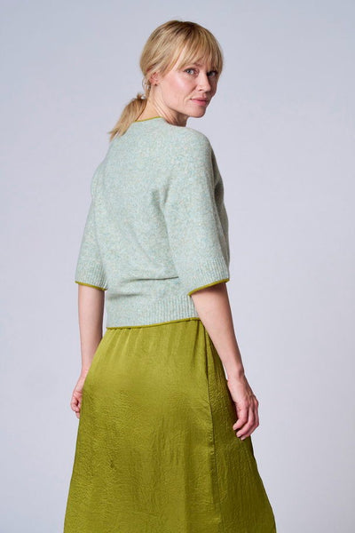 Pull vert de TERRE BLEUE avec manches courtes et bordures contrastées, porté avec une jupe satinée olive.
