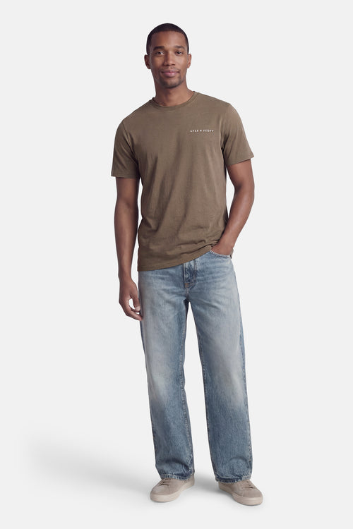 T-shirt kaki à manches courtes de LYLE & SCOTT, porté avec un jean bleu délavé et des chaussures de sport grises.