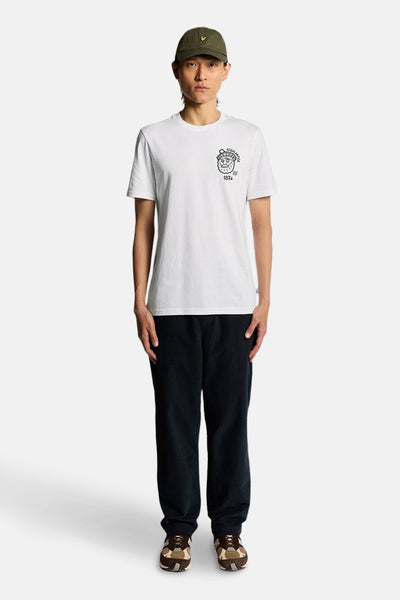 T-shirt à manches courtes blanc de Lyle & Scott, avec un logo imprimé sur la poitrine, porté avec un pantalon et une casquette.