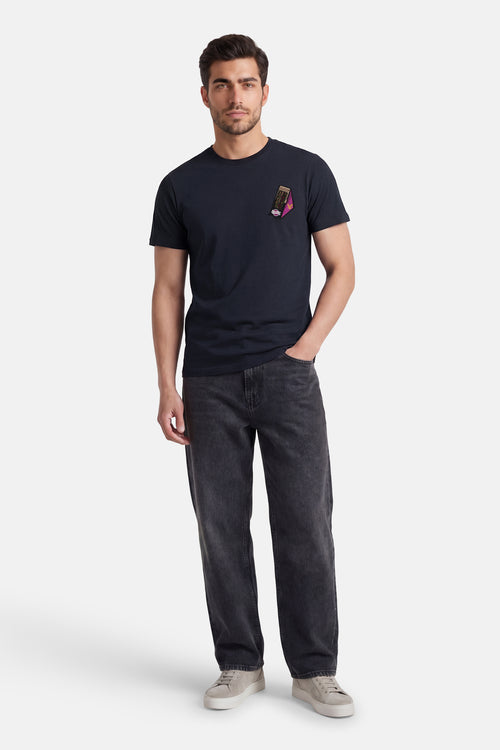 T-shirt bleu à manches courtes de LYLE & SCOTT, avec un écusson décoratif sur le devant, porté avec un jean délavé et des baskets.