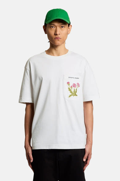 T-shirt blanc à manches courtes de LYLE & SCOTT avec poche poitrine florale rose, porté par un homme en casquette verte et pantalon noir.