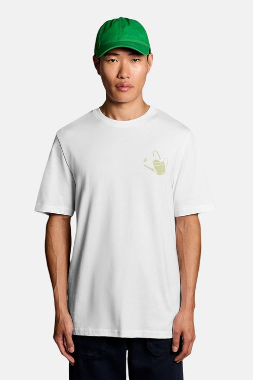 T-shirt blanc de LYLE & SCOTT avec un motif d'arrosoir vert clair, porté par un homme à casquette verte.